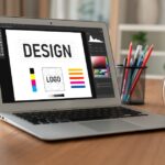 A laptop on a wooden desk displaying a graphic design software interface with the words "DESIGN" and "LOGO," surrounded by color palettes and layout tools.