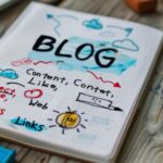 A notebook on a wooden desk with colorful handwritten notes and doodles related to blogging, including words like "BLOG," "Content," "Links," and "Connect," alongside a blue pencil and office supplies.
