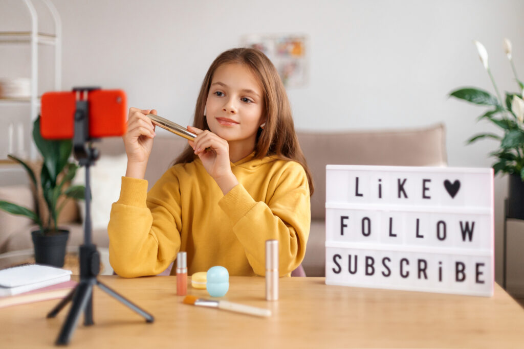 Beautiful young girl shooting video blog about cosmetic A young girl recording Social Media Videos with a smartphone on a tripod, holding a makeup product while sitting at a table with cosmetics and a 'LIKE ♥ FOLLOW SUBSCRIBE' lightbox.
